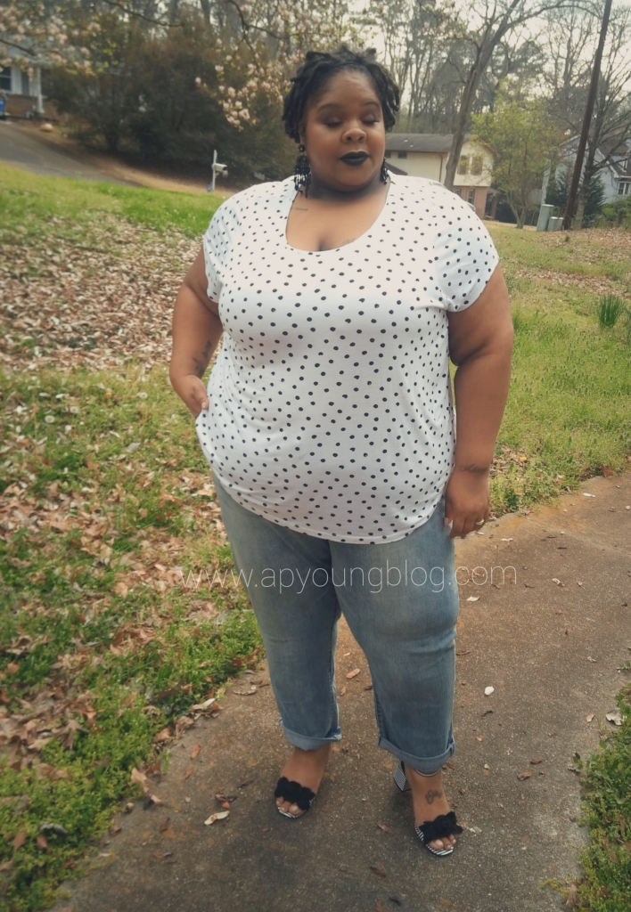 AP an African American Plus size woman is standing outside. Her eyes are slightly closed and she's smiling. She's wearing a white shirt with black polka dots, denim cuffed jeans and a black and white strapped sandal.