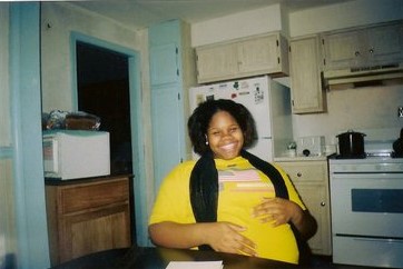 Teen mom, African American seated at a kitchen table holding belly and smiling.